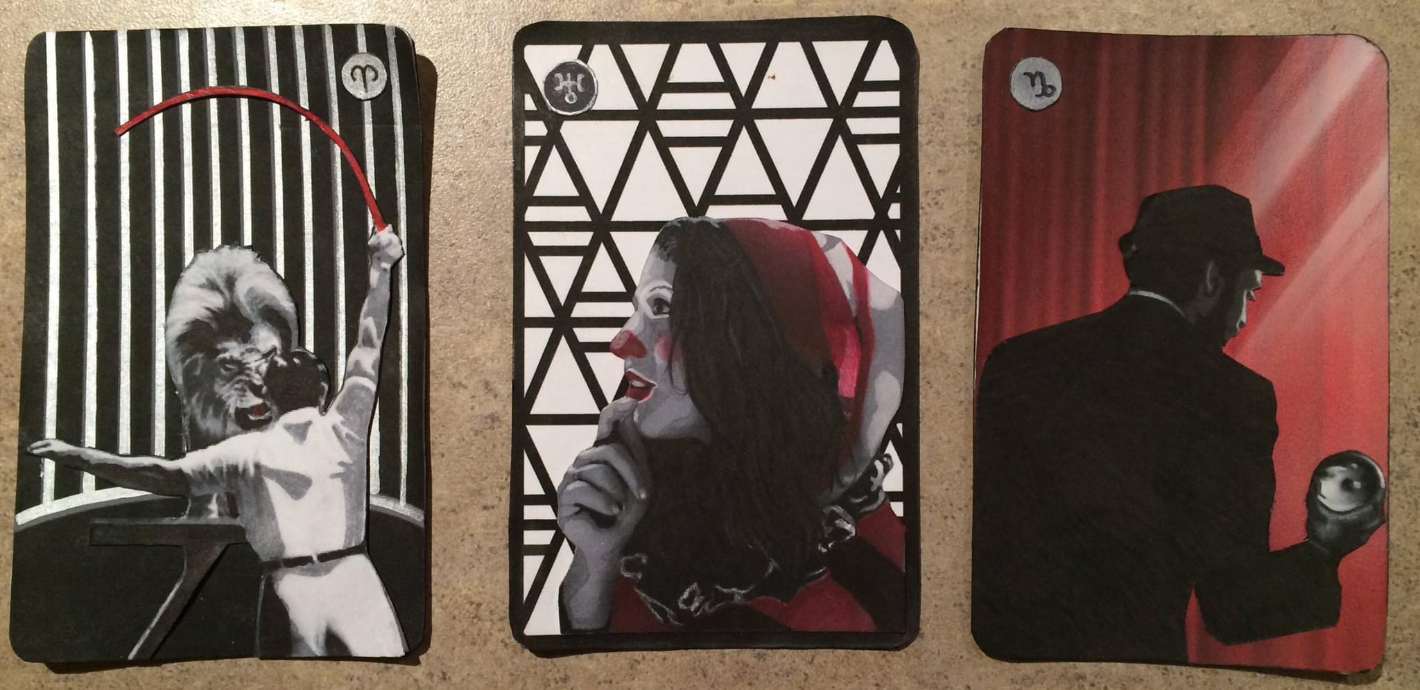 3 cards in a black white and red color scheme, the first is a lion tamer in a cage cracking a red whip at a lion on a platform, the second is a young woman clown with a hand raised thoughtfully to her lips against a triangular backdrop, the third is a man in a suit and bowler hat, his back to the viewer, holding a clear contact juggling/chrystal ball he is backlit behind him is a red curtain there are star sign symbols in the corner of each card