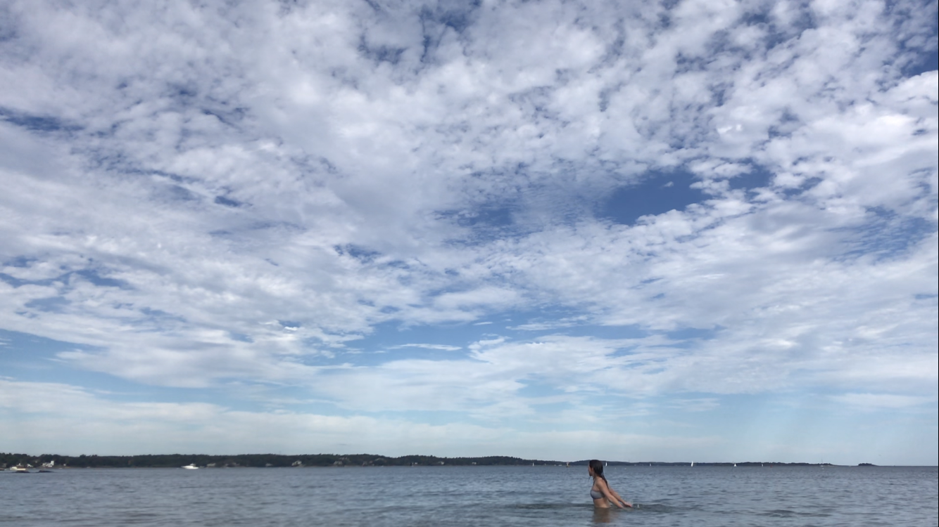 Photo of a woman wading in the ocean. The blue sky is thick with white clouds and dominates the composition