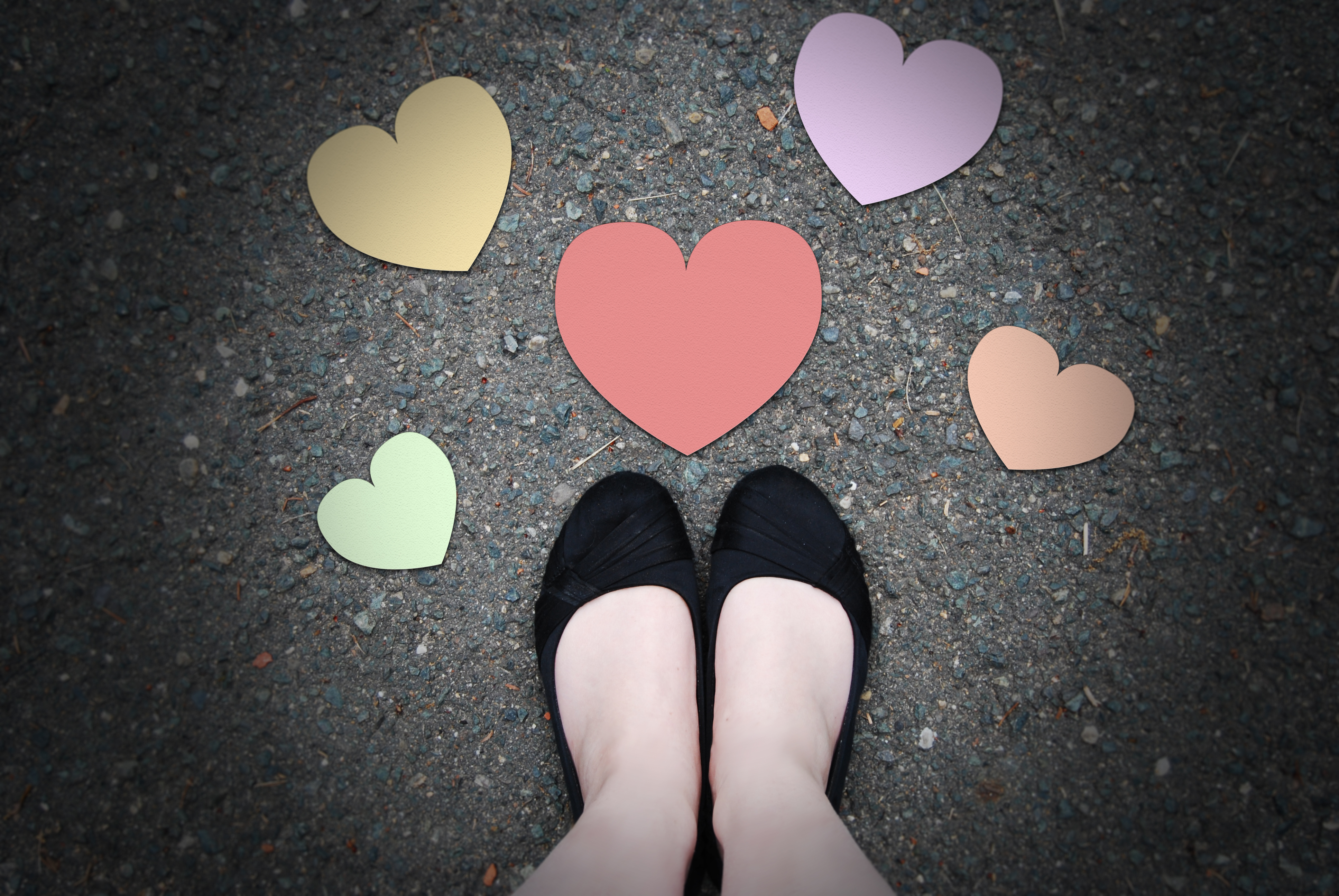 looking down on pavement, at the very bottom edge are a pair of pale white feet in black fabric shoes, around them are colorful paper hearts of varying sizes