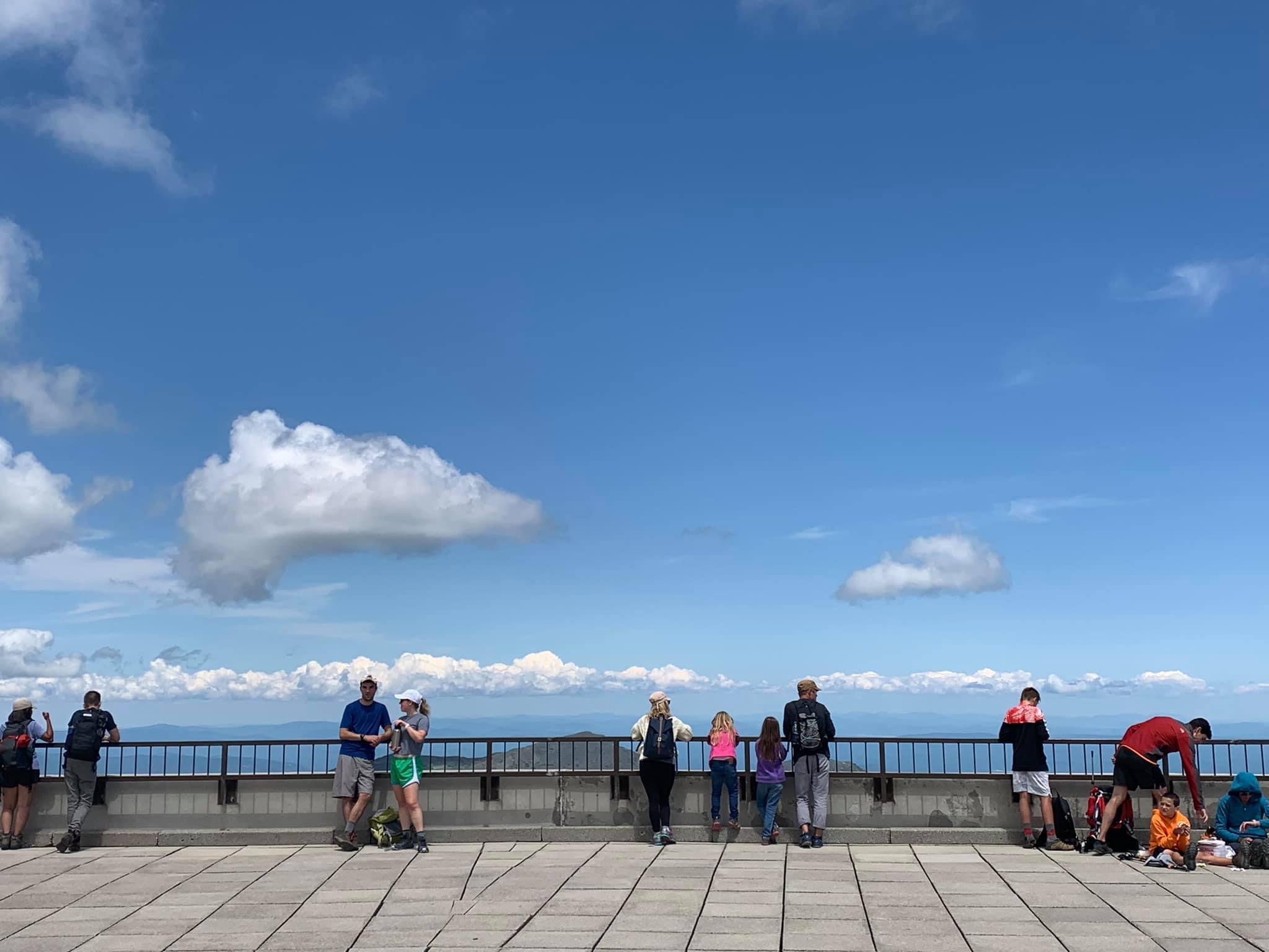 People stand along a railing taking in a view that is mostly blue sky and white puffy clouds.