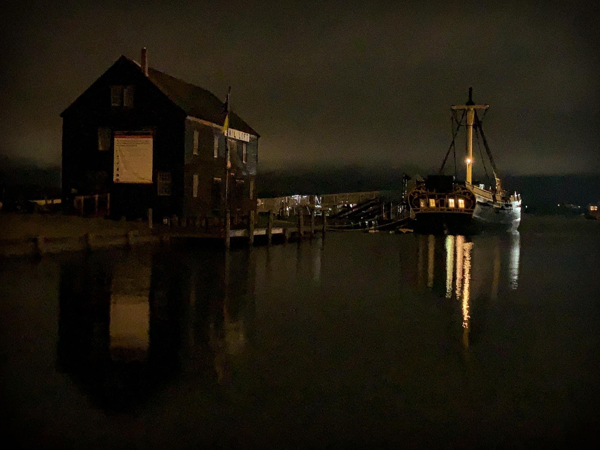 a grainy night time photo of the boat house and The Friendship of Salem docked at night with it's lights reflecting on the still water
