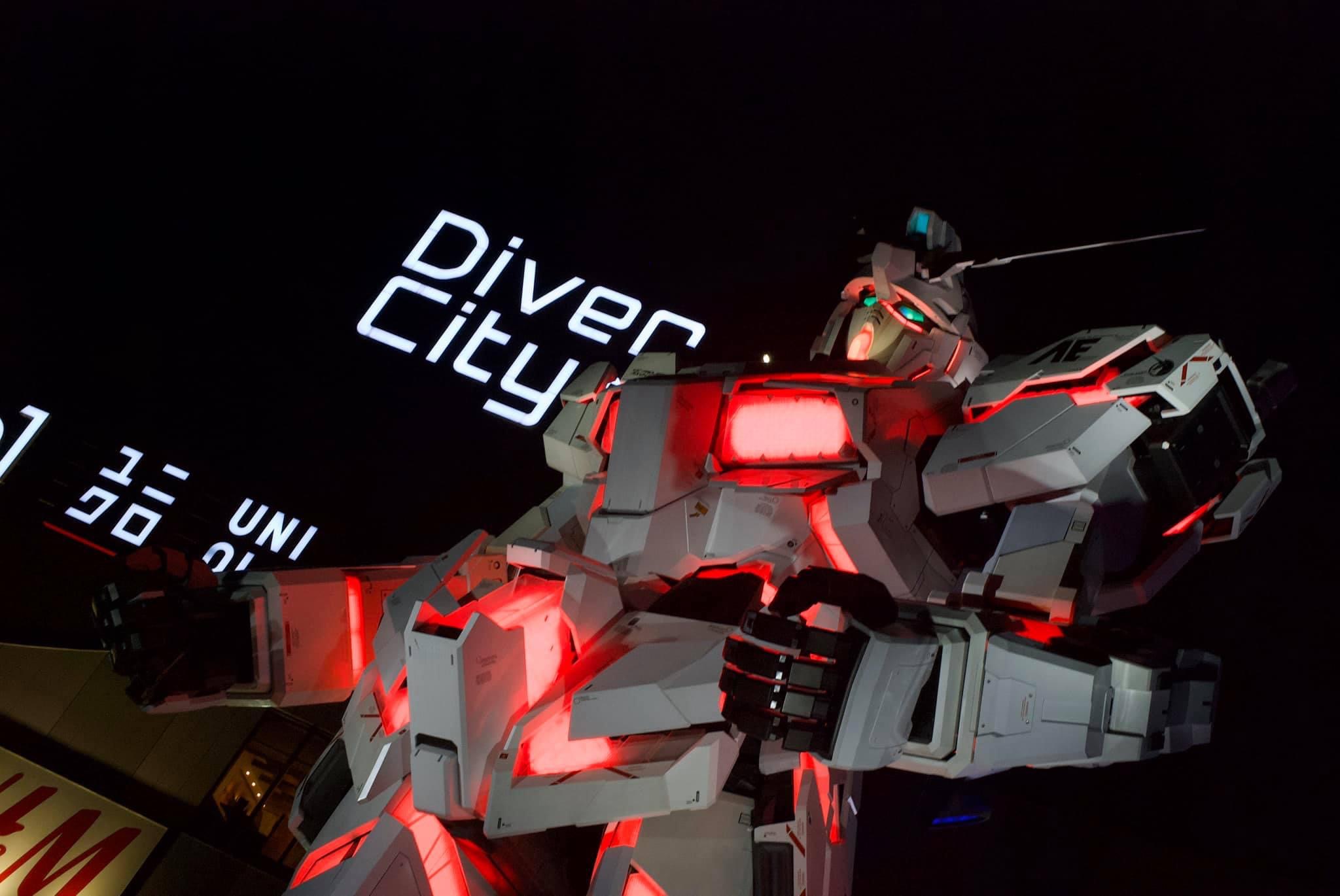A lifesize gundam glowing red against a black night sky, in the background a neon sign for Diver City