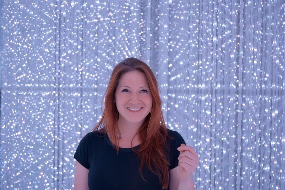 a self portrait of the artist standing and grinning infront of a wall of string lights